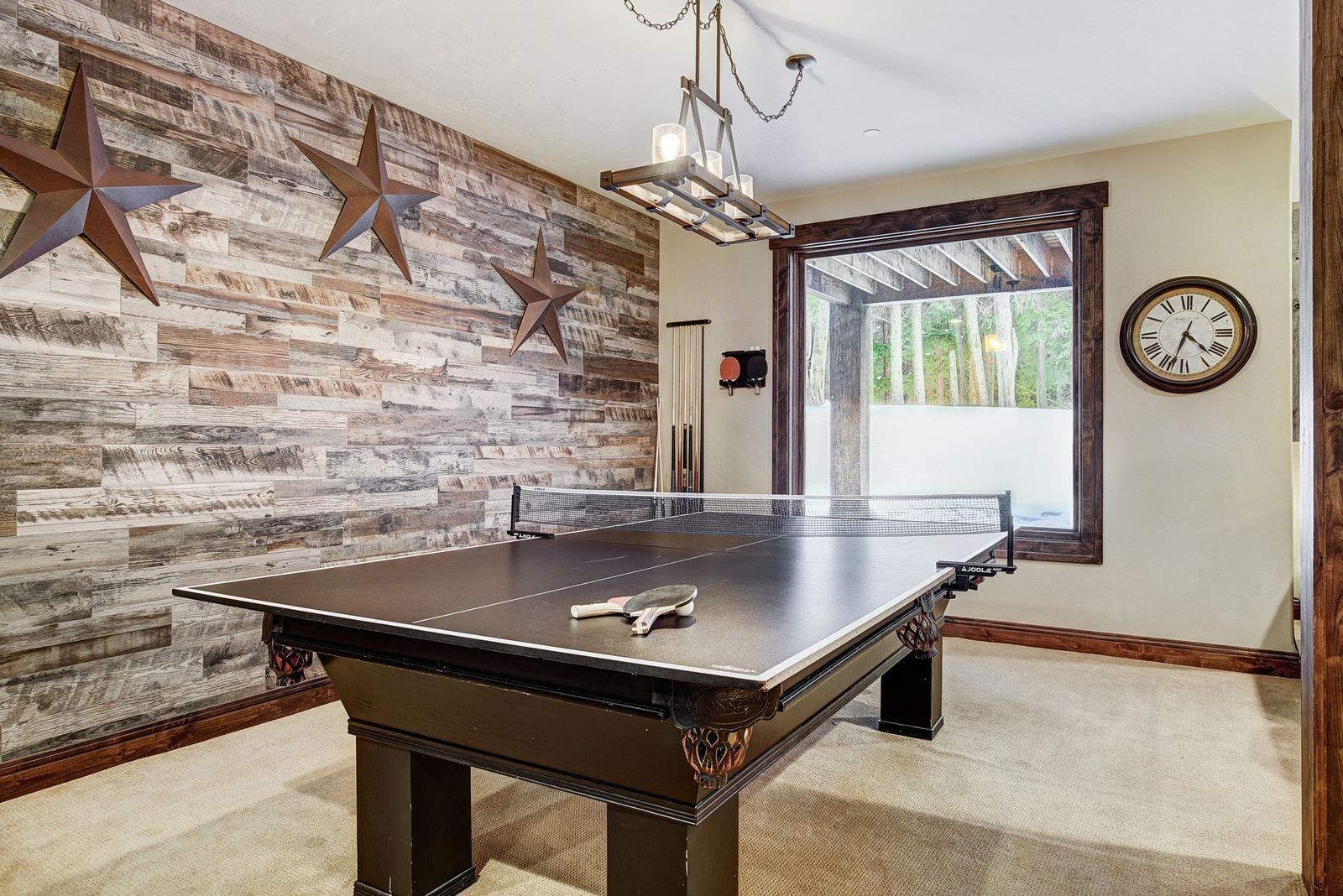 A ping pong table in a room with a rustic wooden wall, a clock, and a window with a view of trees.