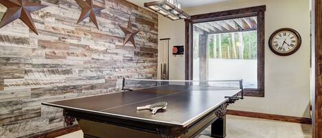 A ping pong table in a room with a rustic wooden wall, a clock, and a window with a view of trees.