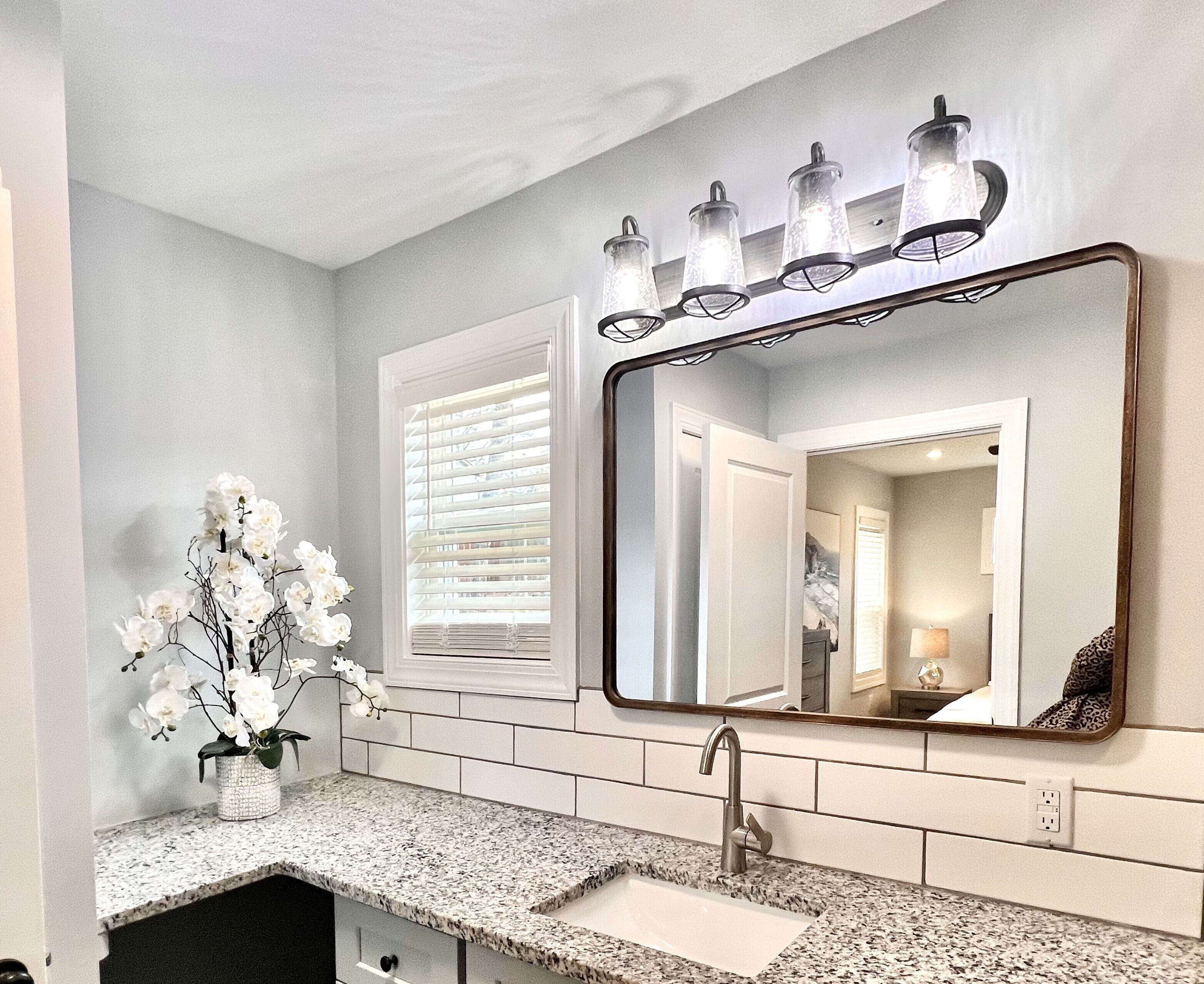 Un baño con encimera de granito, un lavabo, un espejo y un jarrón con flores blancas.