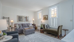 A living room with a grey sofa, a small round table, and a chair. There are two lamps, a potted plant, and a painting on the wall. - 30+ Day 3BR Home One Blk off Historic Main St. Galena w/Pking and W/D (Galena)