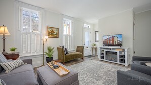 A living room with a sofa, armchair, floor lamp, and a TV on a white entertainment unit. - 30+ Day 3BR Home One Blk off Historic Main St. Galena w/Pking and W/D (Galena)