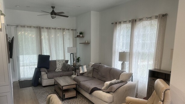Una sala de estar con sofá, sillón, reposapiés, ventilador de techo y televisor sobre un mueble.