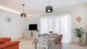 A dining area with a table set for four, surrounded by chairs with cut-out patterns. There is a television on a cabinet, a potted plant, and a large window with sheer curtains. - Large Apartment in the Center of Sorrento, close to Main Transportation Hubs. (Sorrento)