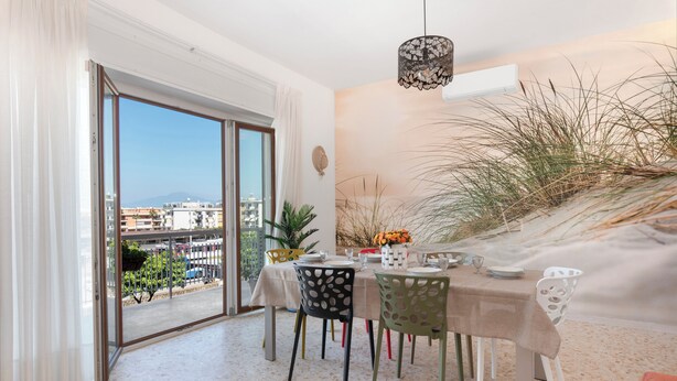 A dining area with a table set for four, a view of the city through a glass door, and a wall mural of a beach scene. - Large Apartment in the Center of Sorrento, close to Main Transportation Hubs. (Sorrento)