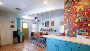 Una cocina compacta con armarios verde azulado, un comedor con sillas naranjas y un mural de pared colorido.