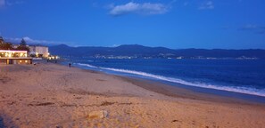 Una playa con un edificio a la izquierda, cielo despejado y montañas a lo lejos.