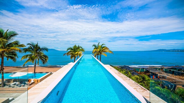 A long swimming pool with clear blue water, surrounded by palm trees and a view of the ocean. - 11th-Floor Oceanview Stay | Infinity Pools + Gym (Puerto Vallarta)