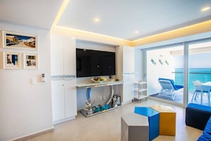 A modern living room with a flat-screen TV, a dining area with a table and chairs, and a balcony with a view of the sea.