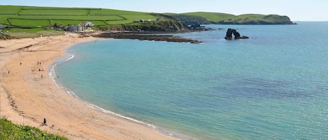Un paisaje costero con una playa arenosa, un promontorio rocoso en el agua y campos verdes al fondo.