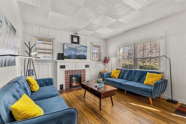 A living room with a fireplace, two blue sofas with yellow cushions, a coffee table, and a TV mounted on the wall.