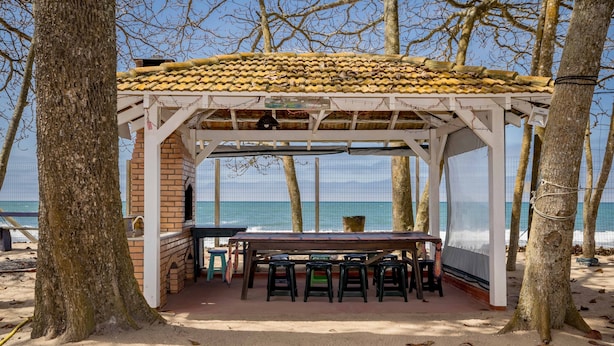 Un kiosque au bord de la plage, doté d’un toit en tuiles, d’une table et de plusieurs chaises.