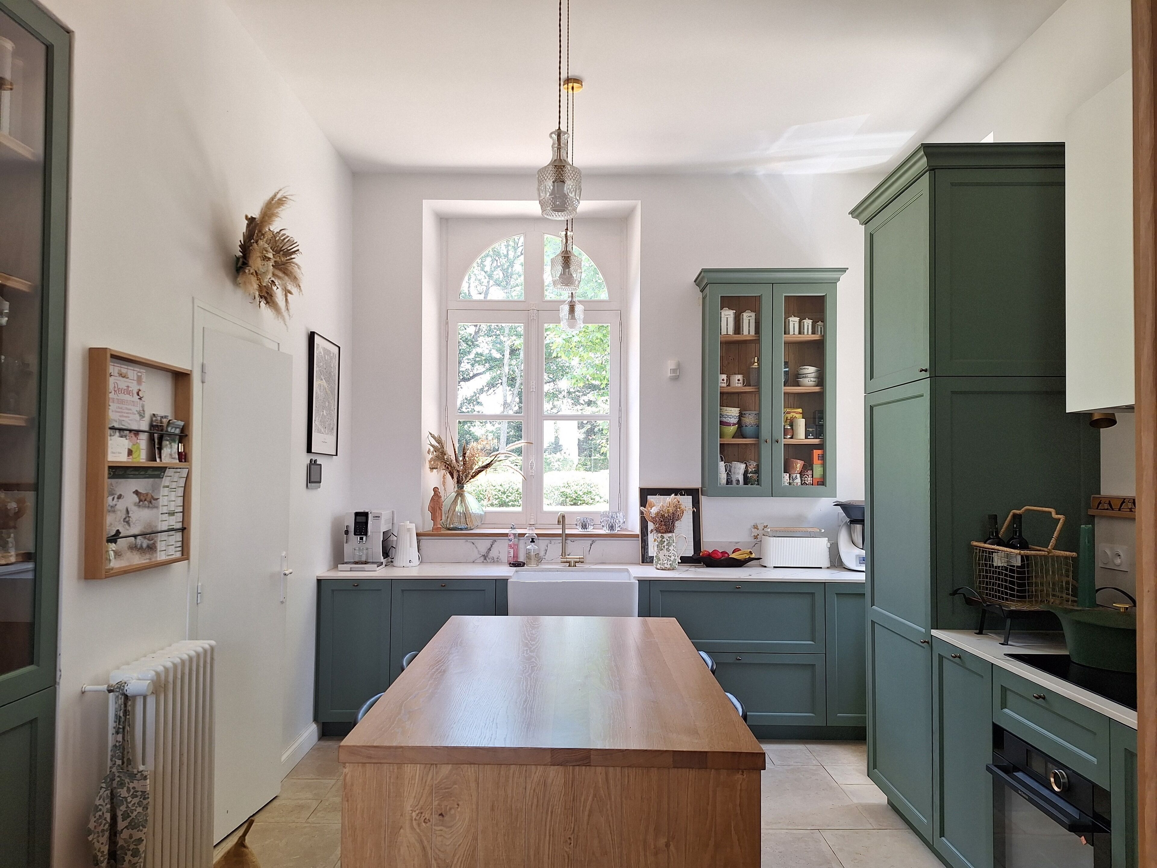 A kitchen with a central island, green cabinets, and a window with a view of greenery.