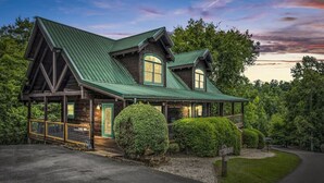 A two-story log cabin with a green metal roof, a covered porch, and a well-maintained lawn. - Near Attractions-Pool Table-Hot Tub-Fire Pit (Pigeon Forge)