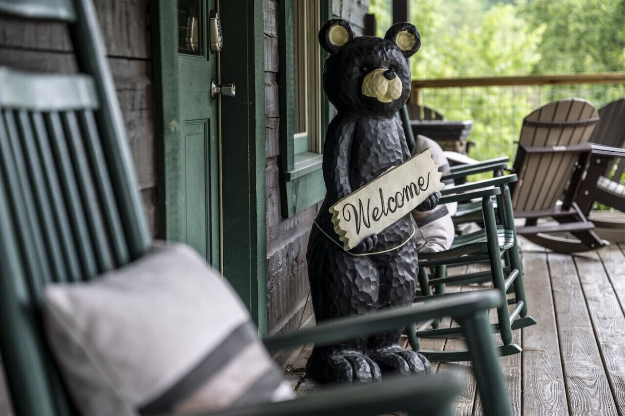 Eine hölzerne Veranda mit Schaukelstühlen und einer schwarzen Bärenstatue, die ein 'Welcome'-Schild hält.