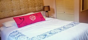 A neatly made bed with a white and blue patterned bedspread, a red pillow with a floral design, a bedside lamp, and a patterned wall.