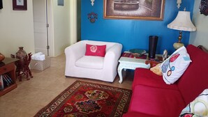 A living room with a white armchair, a red sofa, a patterned rug, a small table, and a painting on the wall. - Private Garden Villa in Manchester's Cool Highlands  (Cross keys)