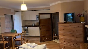 Quarto compacto com mesa de jantar de madeira, cadeiras, cama, cômoda de madeira, televisão e cozinha equipada com armários e micro-ondas.