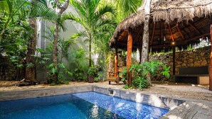 Une piscine dont le bord est revêtu de carreaux, entourée de plantes tropicales et bordée d’une structure à toit de chaume.
