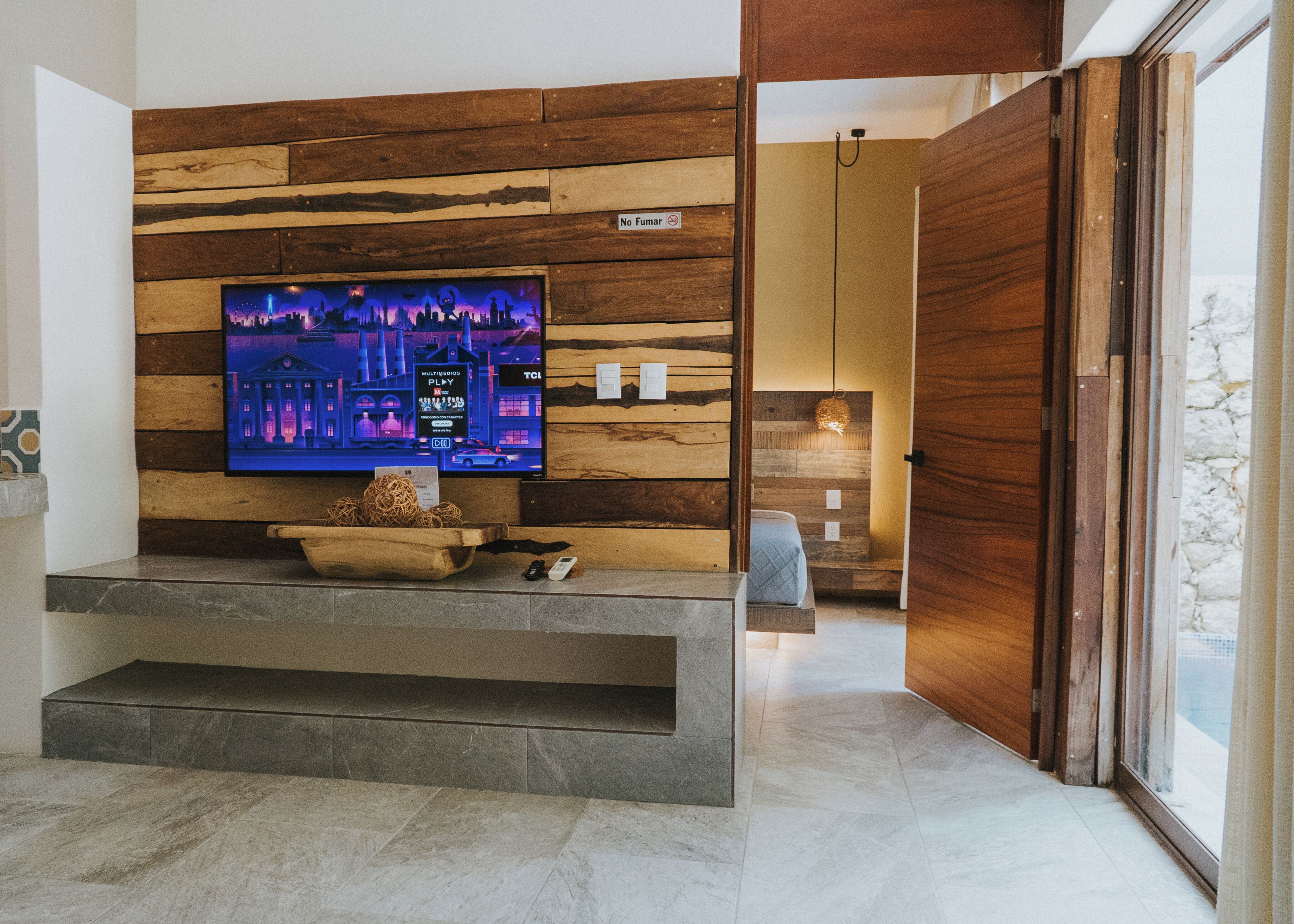 A modern living room with a flat-screen TV mounted on a wooden wall, a stone bench, and a view of a cityscape.