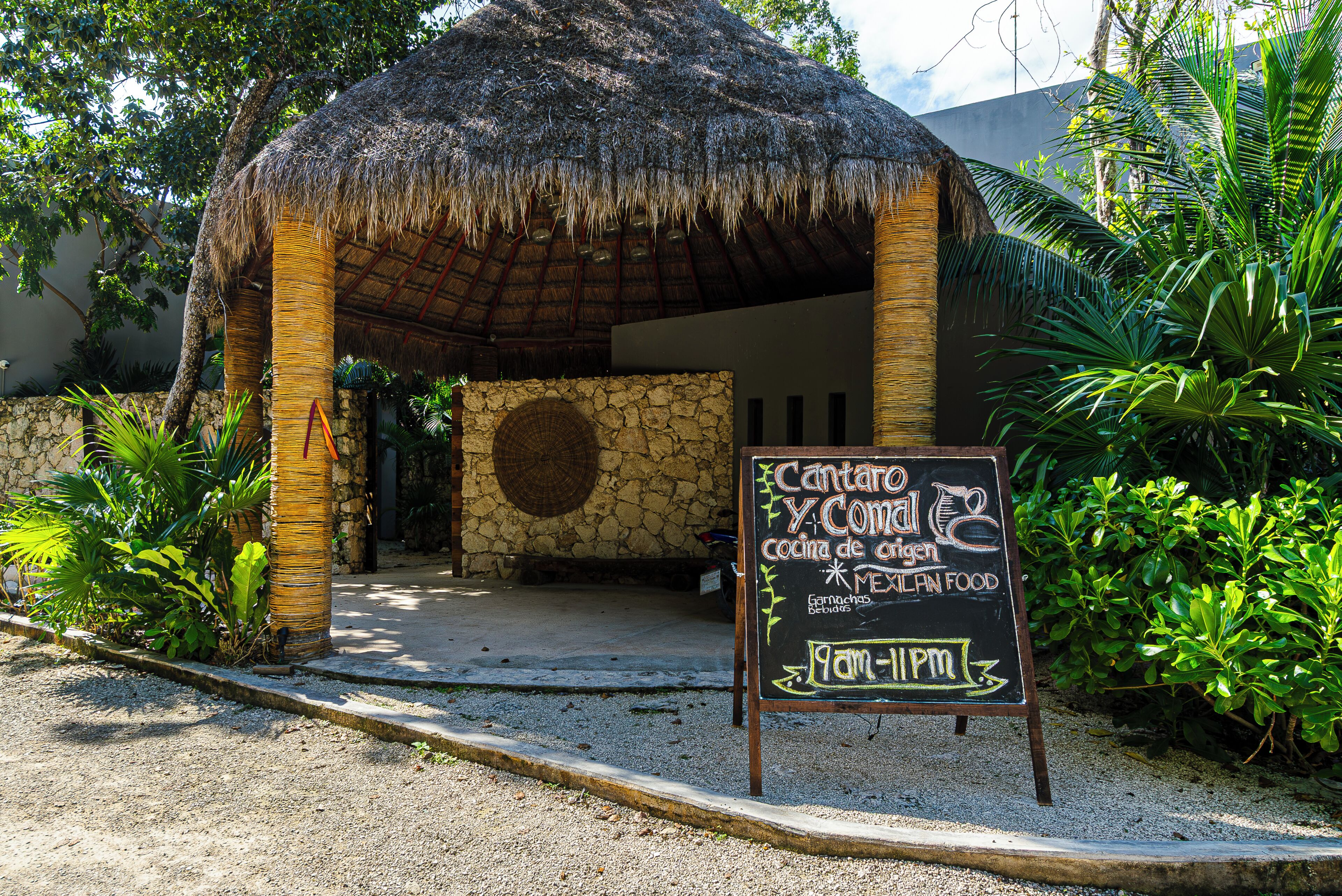 A thatched-roof structure with a stone wall and a signboard advertising Cantaro Y Cocina de Cocina Mexican food.