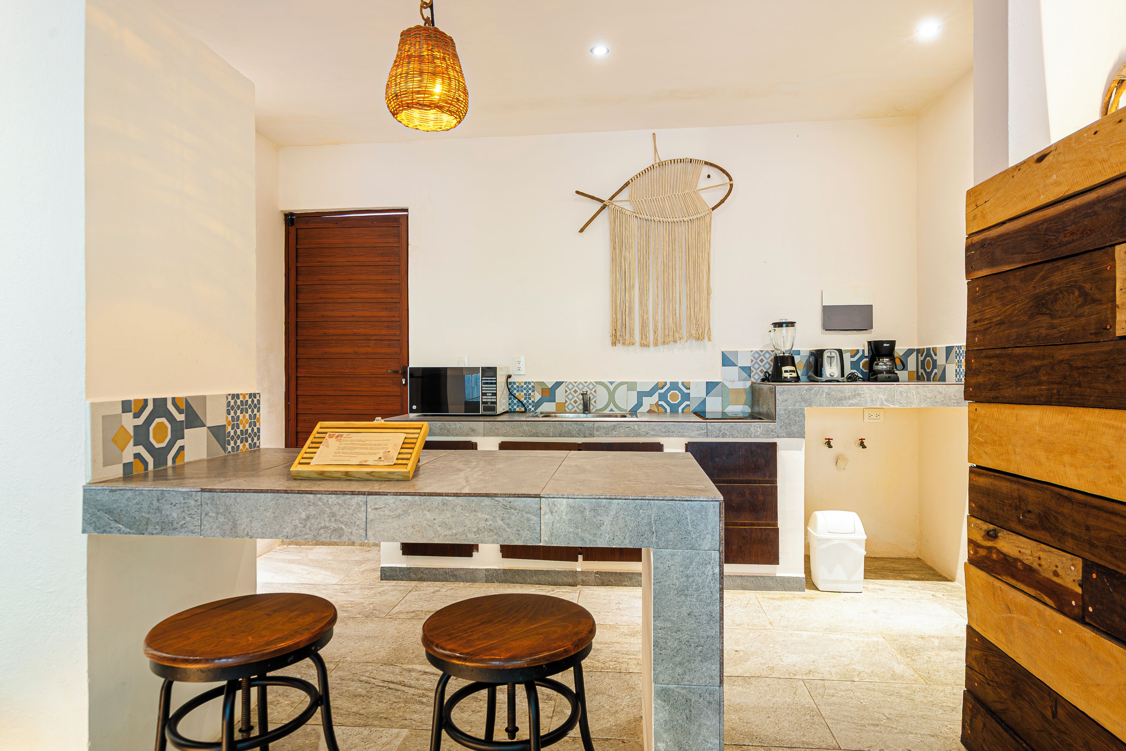 A kitchen with a wooden door, a hanging macrame wall decoration, a coffee machine, and a trash bin.