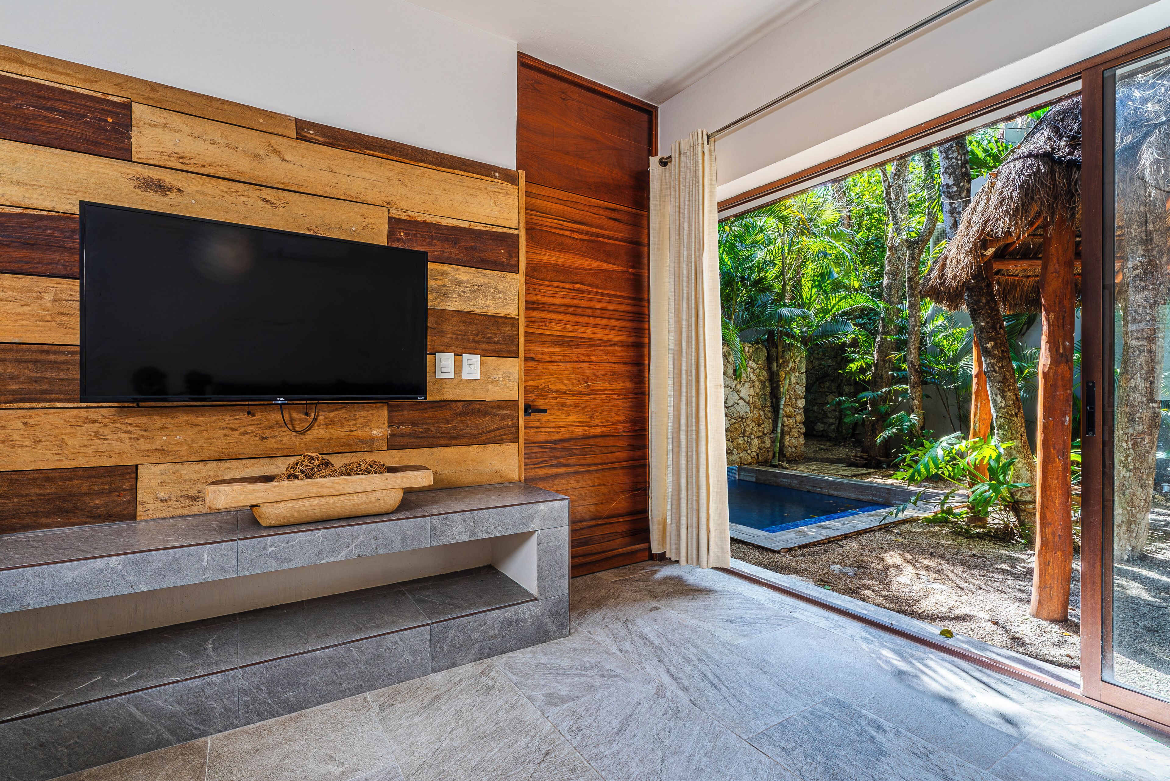 A modern living room with a flat-screen TV, a stone bench, and a glass door leading to a pool area.