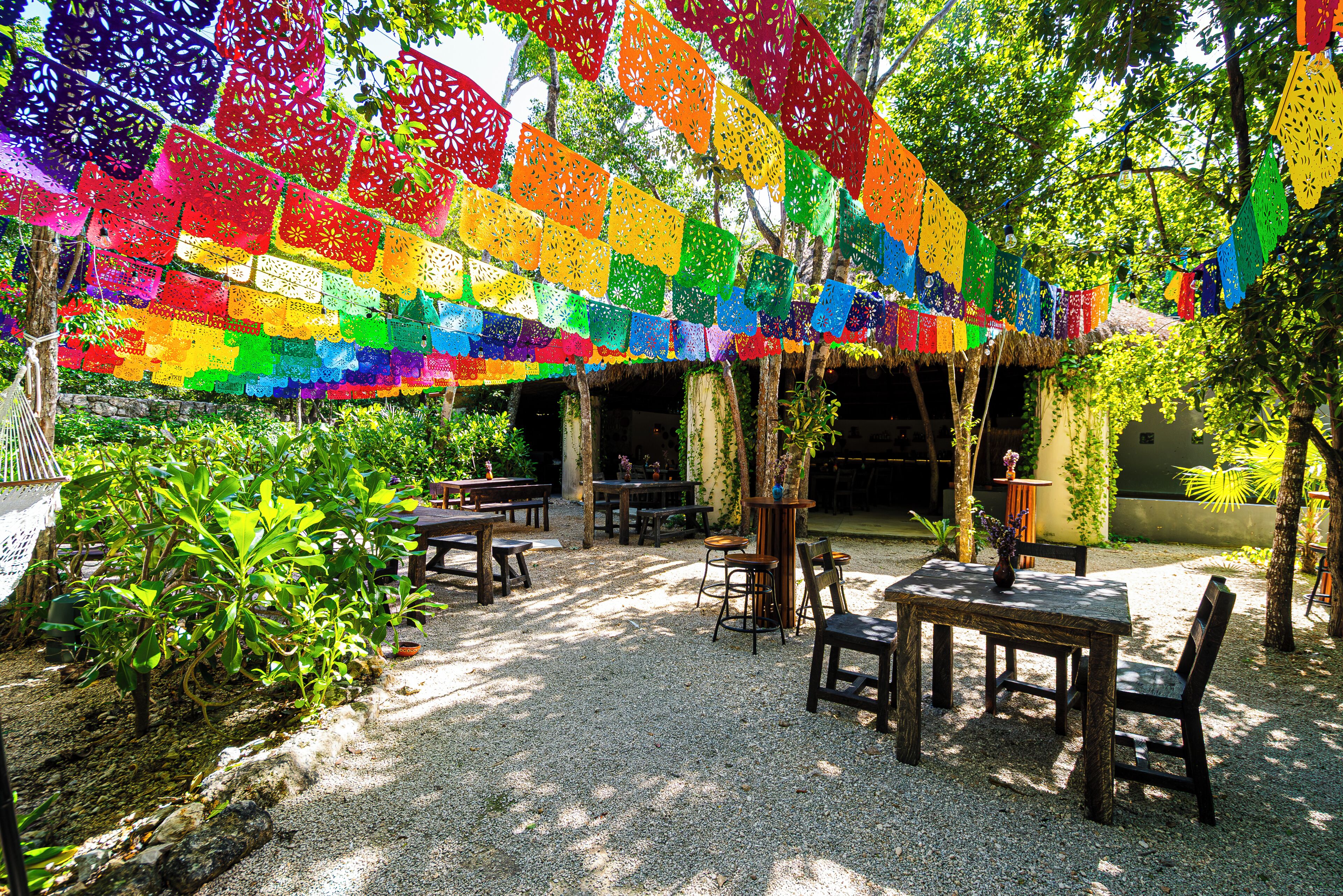 Un espace extérieur aménagé en terrasse, avec des tables et des chaises, entouré d’arbres et agrémenté d’une décoration colorée suspendue.