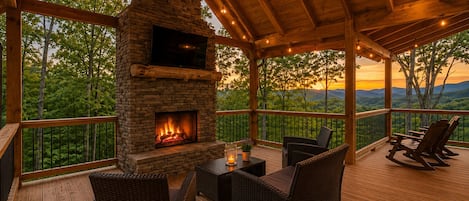 A covered outdoor deck with a stone fireplace, wicker furniture, and a view of the landscape.