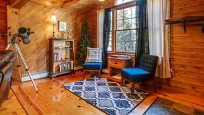 A cozy cabin interior with wooden walls, a telescope, a bookshelf, a blue armchair, and a large window with curtains. - The Rexford Cabin: Easy Drive to PA Grand Canyon! (Gaines Township)
