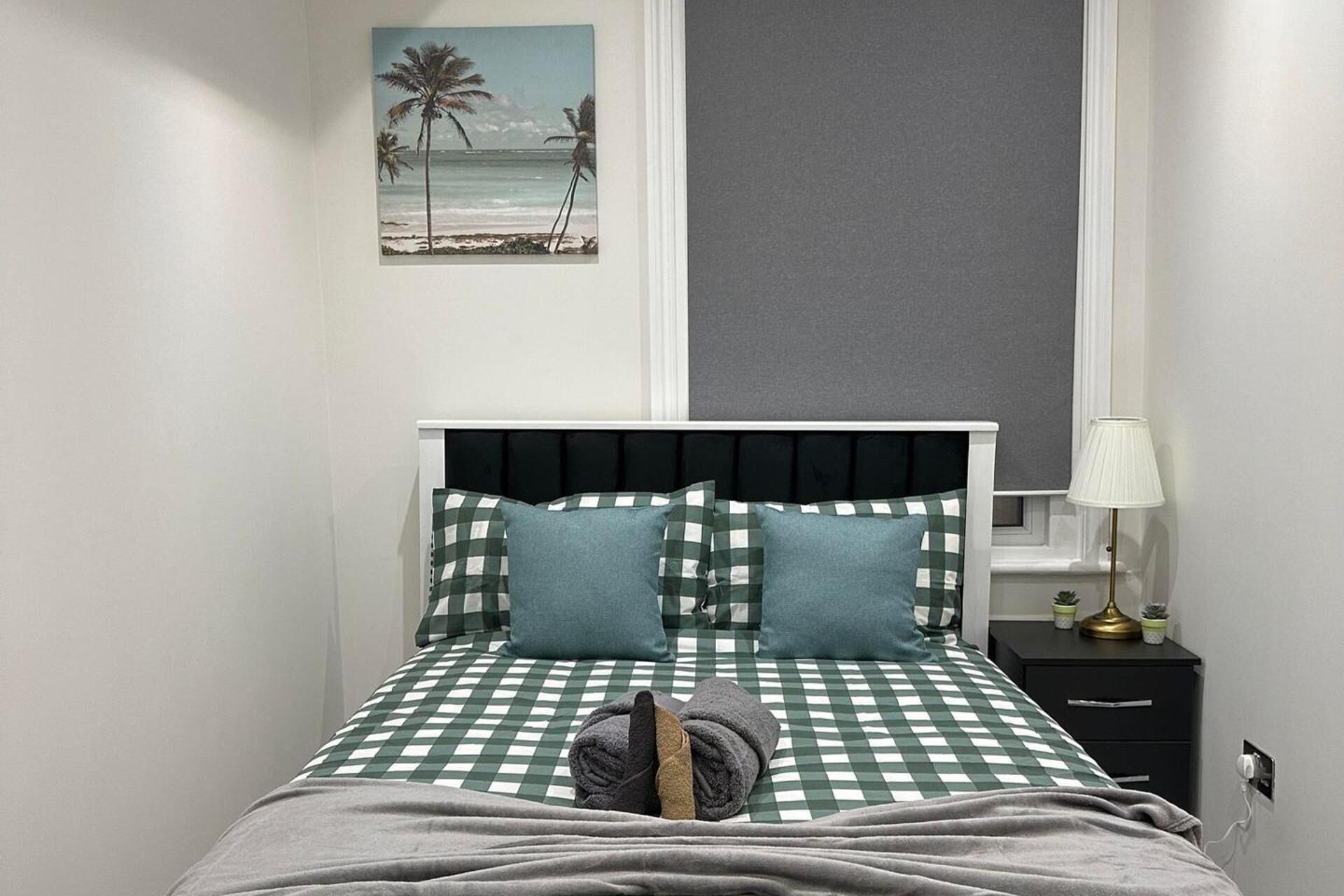 A neatly made bed with checkered bedding, a bedside lamp, and a small bedside table with a lamp and two potted plants.