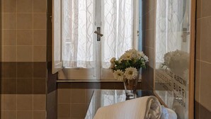 A bathroom with a glass shower enclosure, a vanity with a mirror, and a window with decorative curtains. - Luxury suite off Piazza del Popolo (Roma)