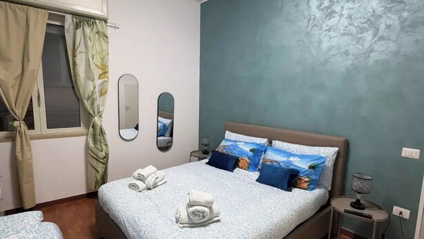 A bedroom with a bed, two bedside tables, a window with curtains, a mirror, and a wall with a textured pattern.