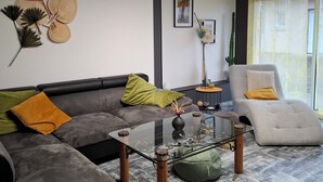 Un salon moderne avec un canapé d’angle, une table basse en verre, un fauteuil blanc et une grande fenêtre ornée de rideaux légers.
