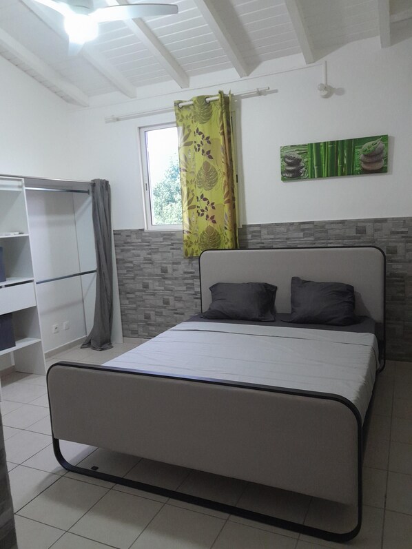 A bedroom with a bed, a wardrobe, a window with a curtain, and a wall with a decorative piece. - A LA Campagne : Haut DE Villa Equipee, Spacieux, Tout Confort (Lamentin)