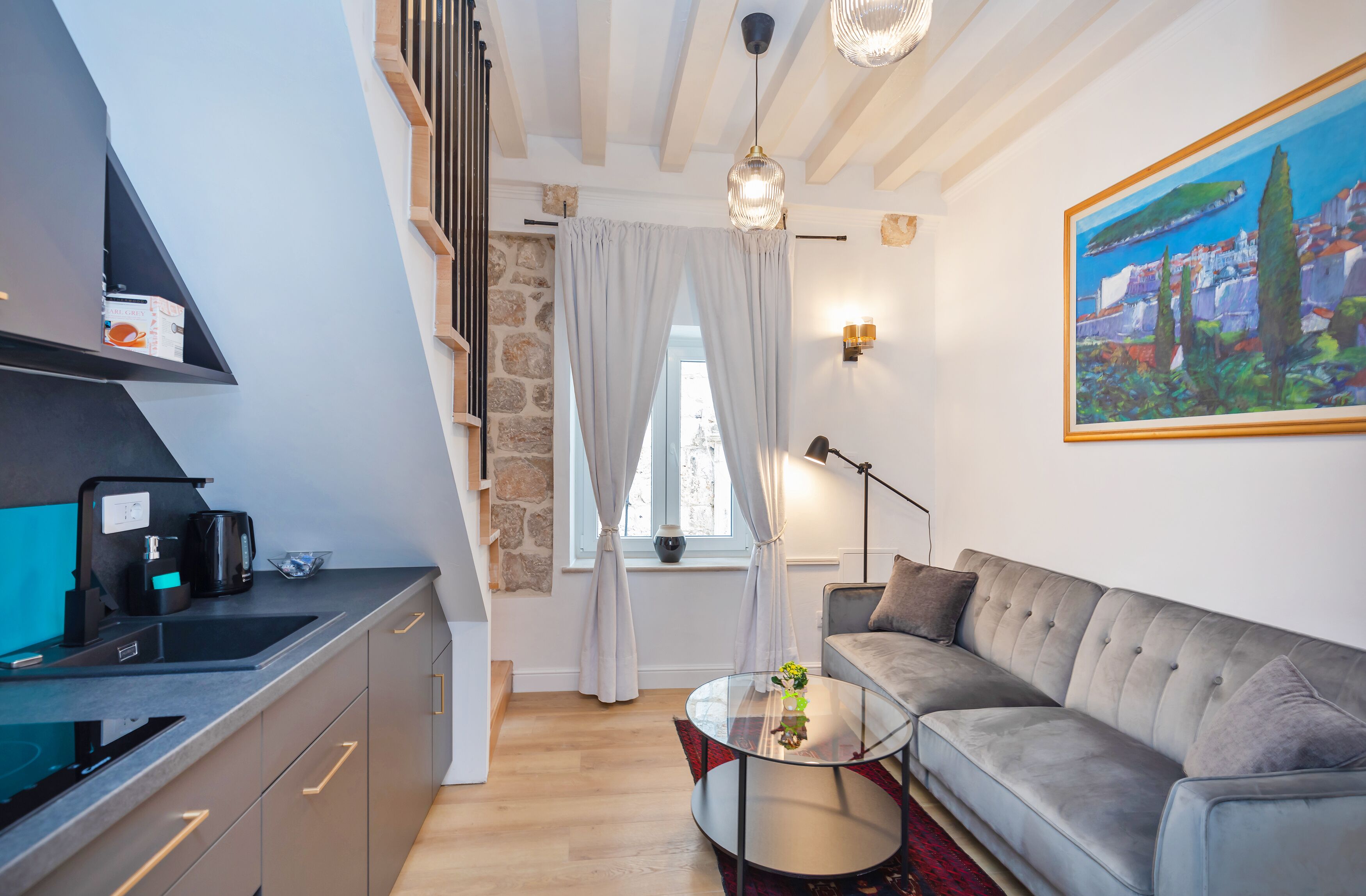 Eine moderne Küche mit Einbauofen, Kaffeemaschine und Spüle. Ein graues Sofa mit Kissen und ein Gemälde einer Küstenlandschaft an der Wand.