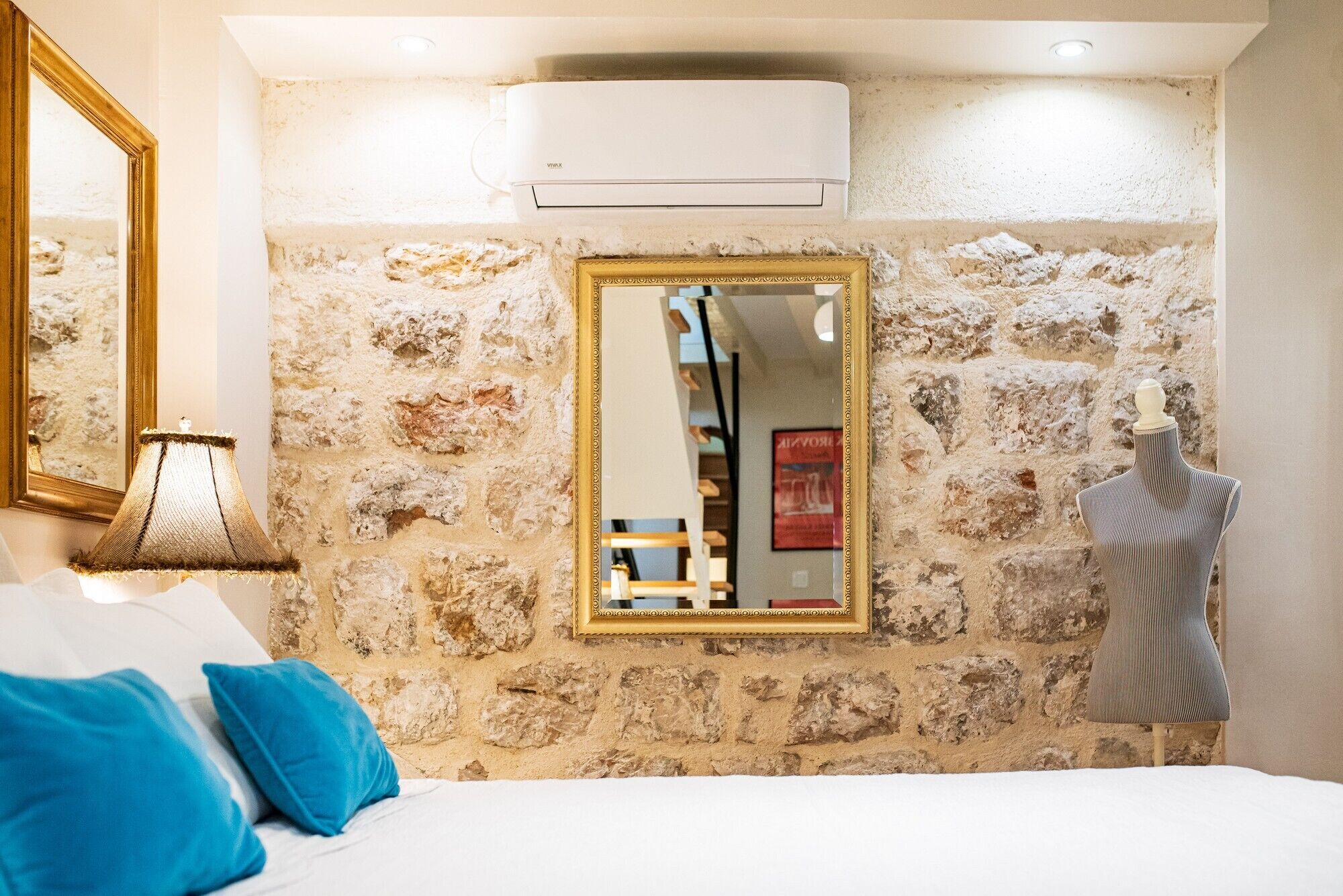 A bedroom with a stone wall, a large mirror, a bed with blue pillows, and a mannequin.