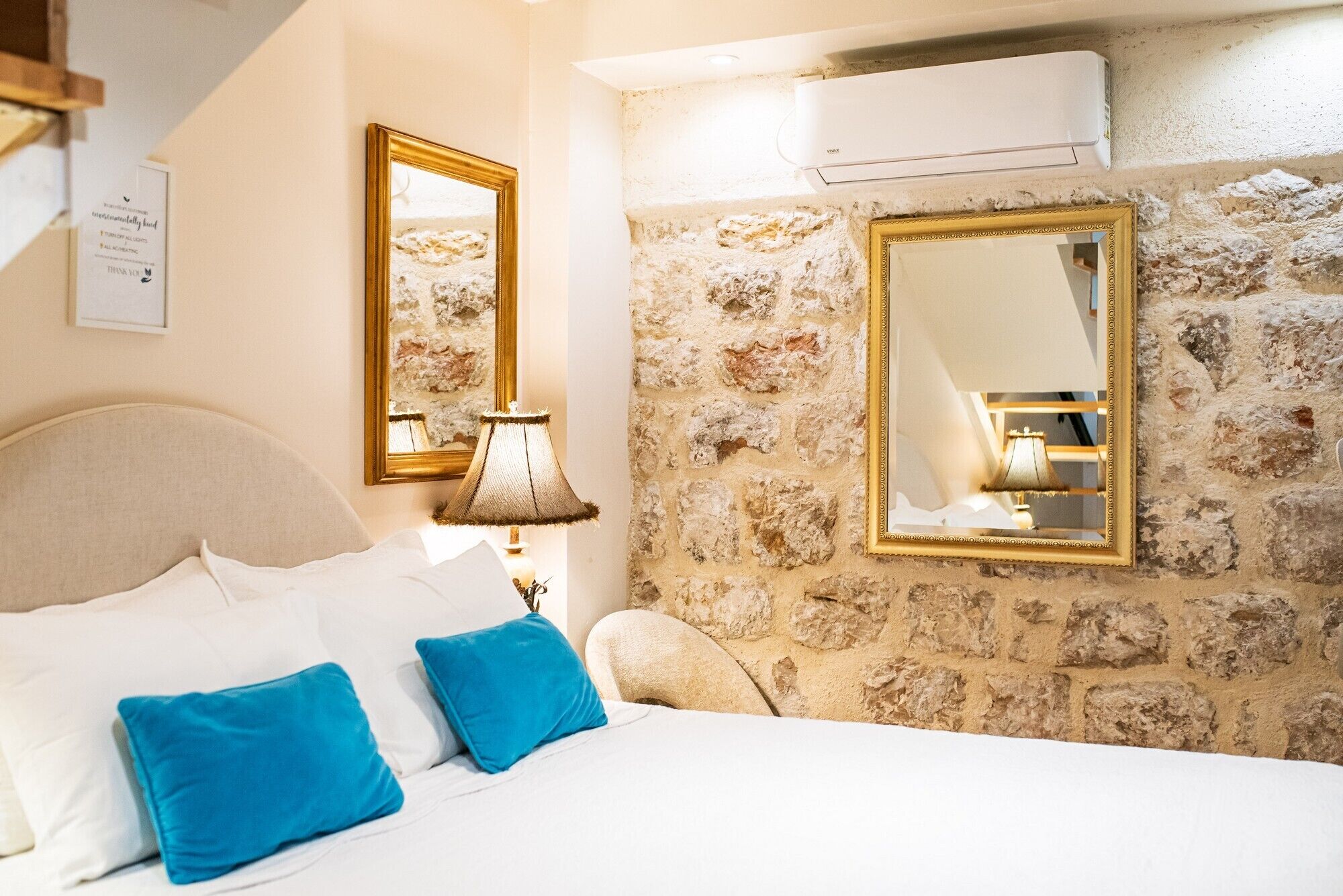 A bedroom with a stone wall, a bed with white and blue pillows, a lamp, and two mirrors with gold frames.