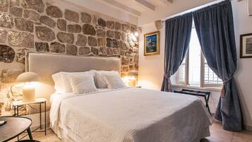 Une chambre avec un mur en pierre, un lit avec une literie blanche, une table de chevet avec une lampe et une fenêtre avec des rideaux.