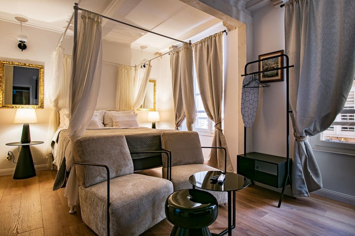 A four-poster canopy bed with a view of the city, a small round table, a chair, and a floor lamp.