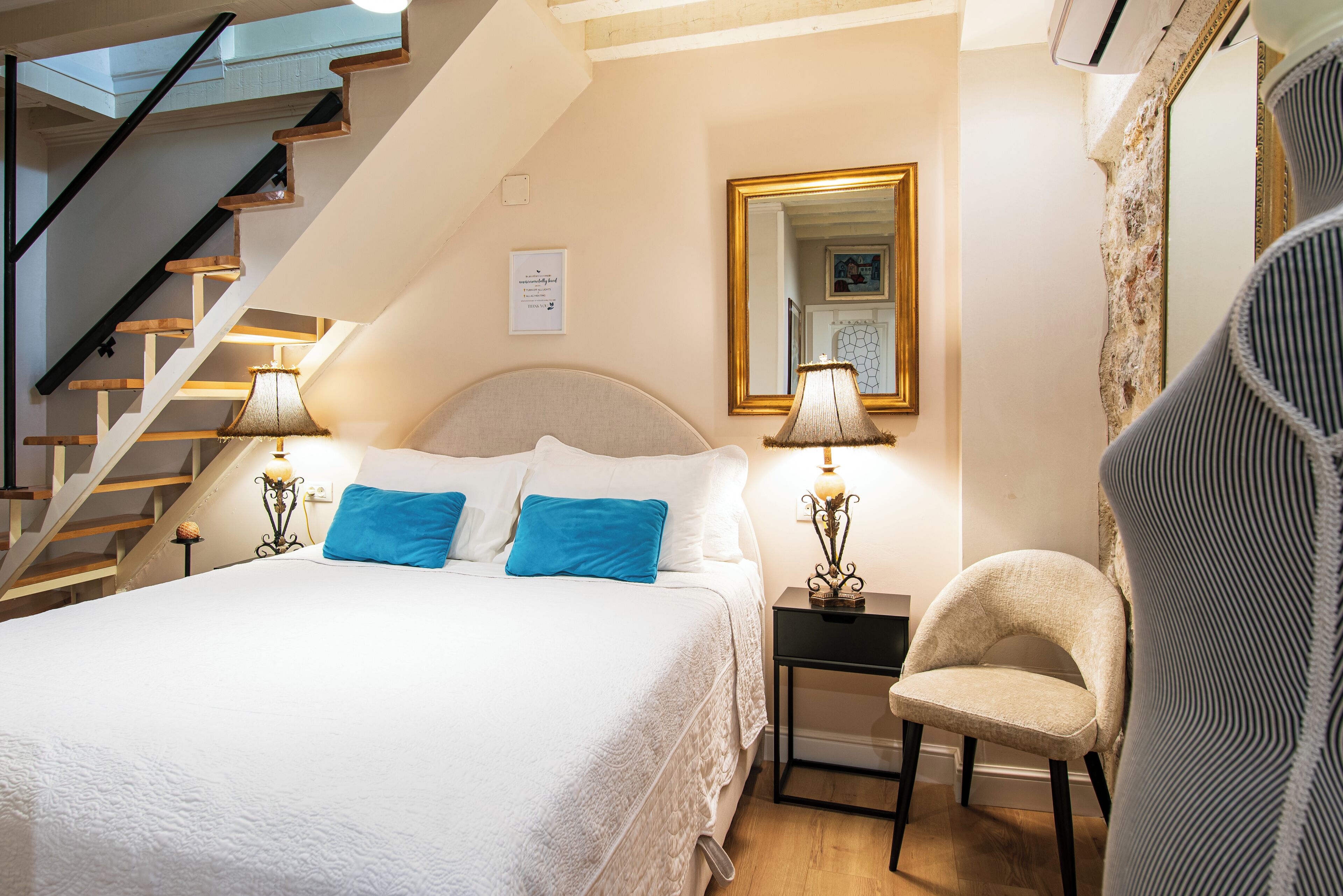A bedroom with a bed, two bedside lamps, a chair, a mirror, and a staircase.