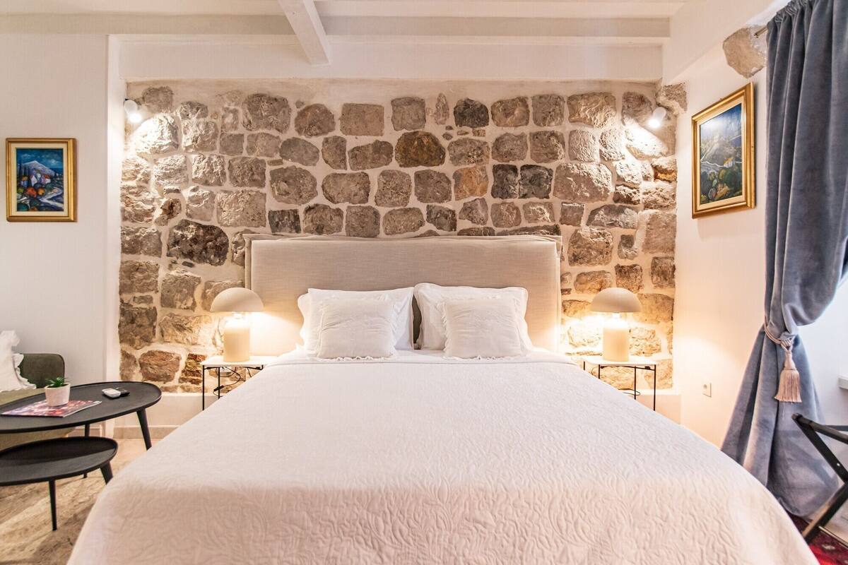 A bedroom with a stone wall, a large bed with white bedding, two bedside lamps, and a small table with a plant.