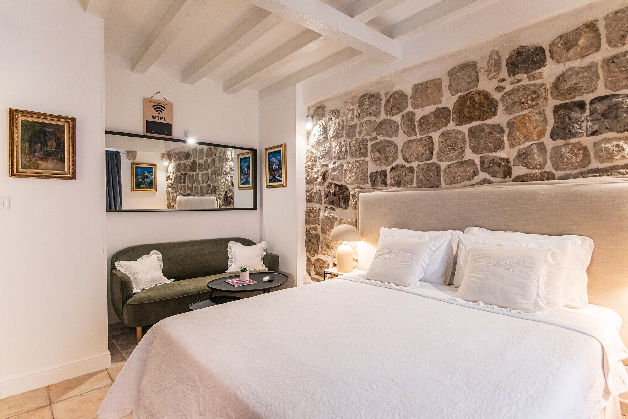 Ein Schlafzimmer mit Steinwand, einem Bett mit weißer Bettwäsche, einem grünen Sofa, einem kleinen Tisch und einem Spiegel mit Kunstwerken.