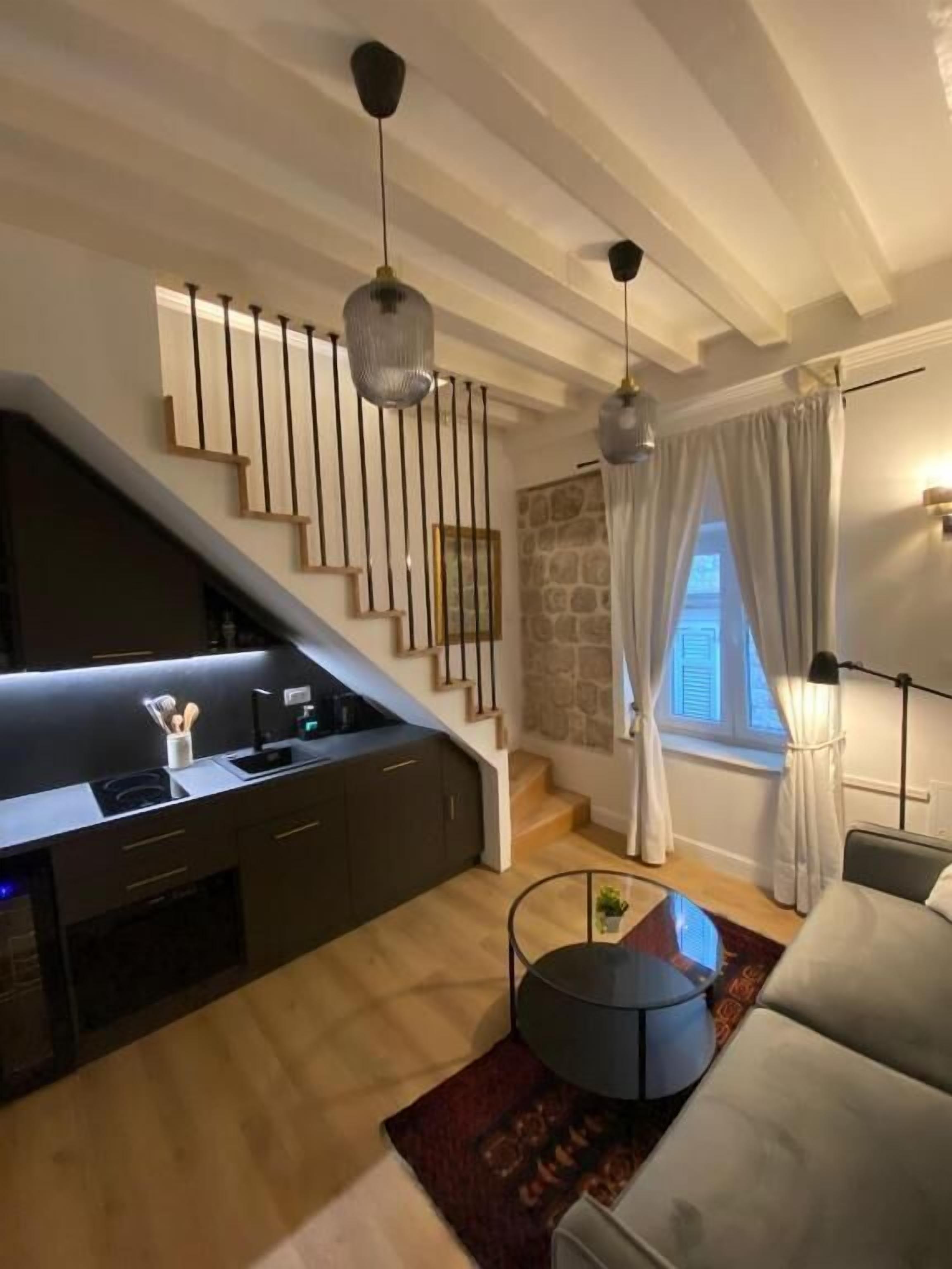 A modern living room with a kitchen area, a staircase, and a large window with curtains.