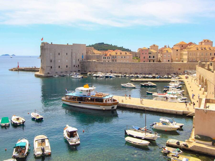 A harbor with boats docked, a large building with a flag, and a stone wall.