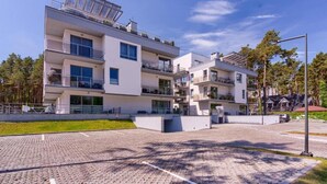 Un edificio residencial moderno de varios pisos con balcones, un camino de entrada pavimentado y vegetación circundante.