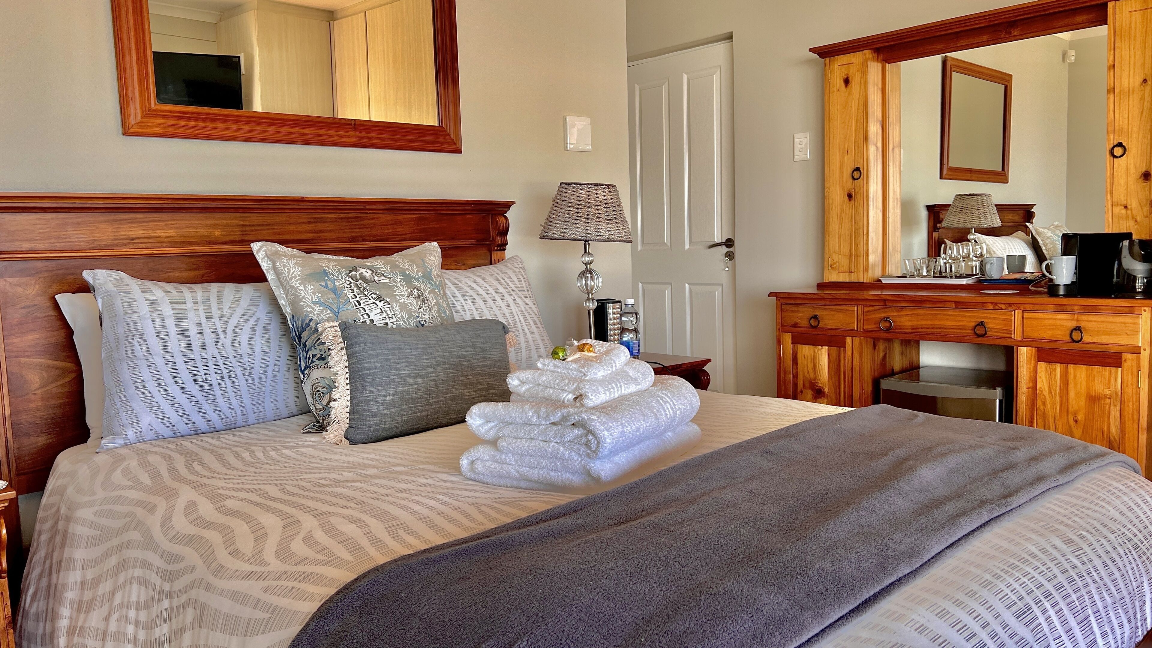 A neatly made bed with patterned bedding, a bedside lamp, and a wooden dresser with a mirror.