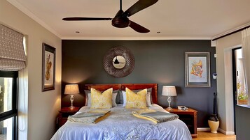 Un dormitorio con una cama, dos mesitas de noche, un ventilador de techo y un espejo decorativo grande en la pared.