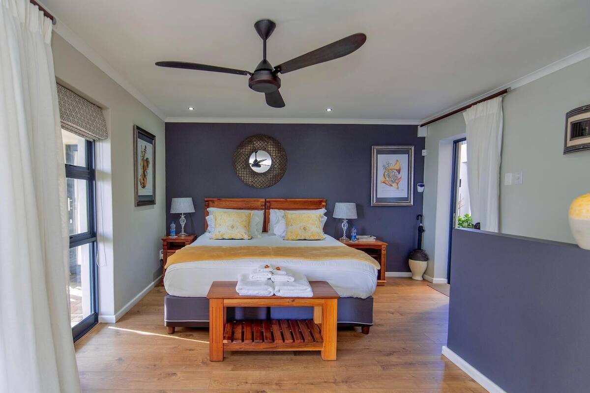 Une chambre bien éclairée, dotée d’un grand lit, d’un ventilateur de plafond et d’une table de chevet en bois sur laquelle sont posées des serviettes.