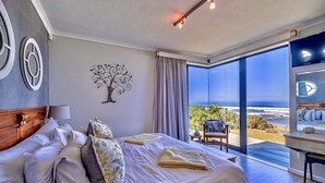 Un dormitorio con una cama grande, un cuadro de un árbol, vistas al océano y un balcón con una silla.