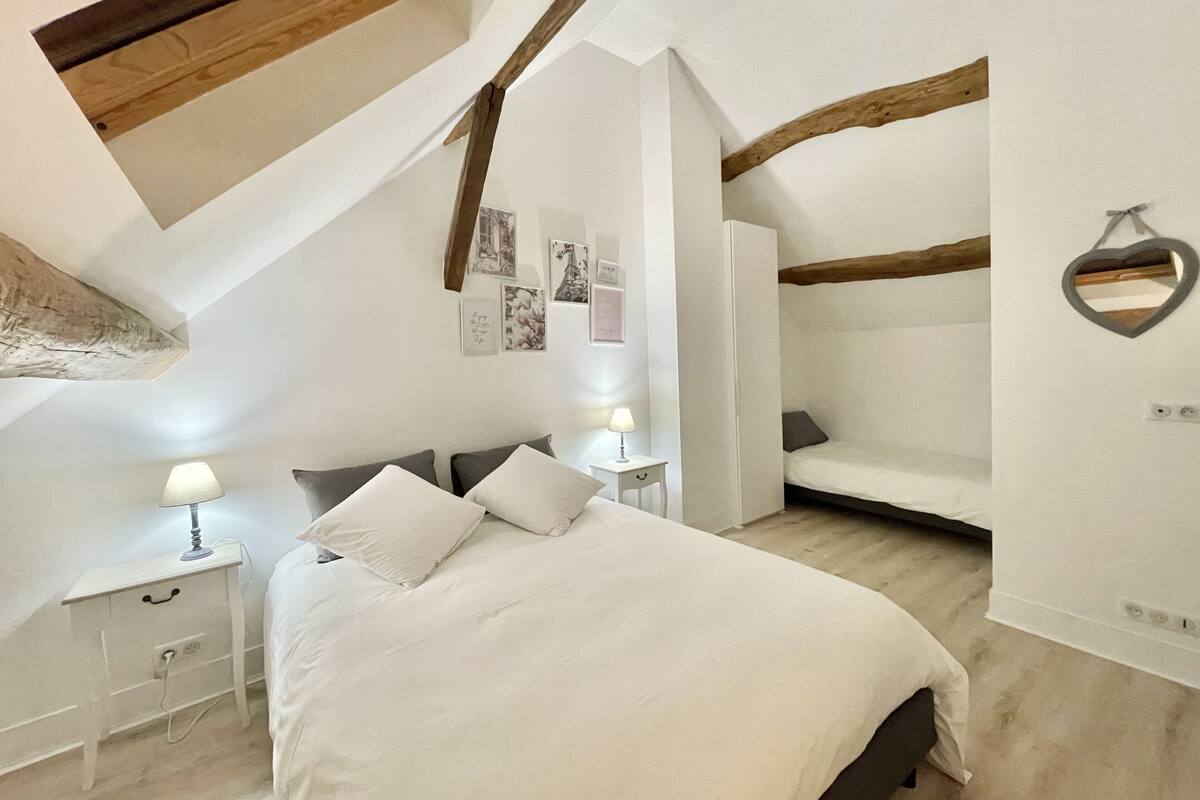 A bedroom with a sloped ceiling, exposed wooden beams, a bed with white bedding, a bedside table with a lamp, and a small nightstand.